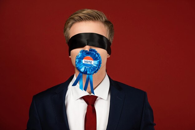 Foto man imiteert met blinddoek op ogen en badge met stembelettering in mond op rode achtergrond