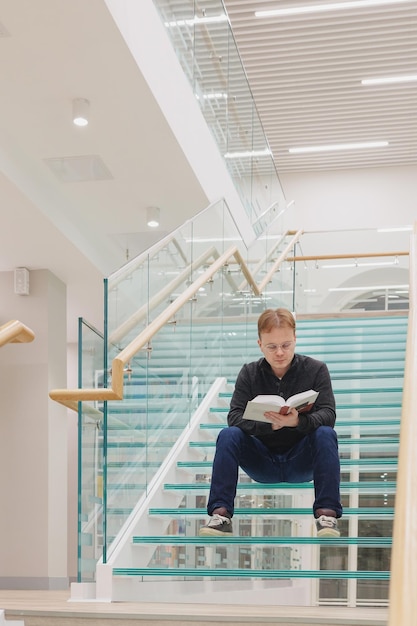 Man in bibliotheek leest boek zittend op trappen van glazen trap