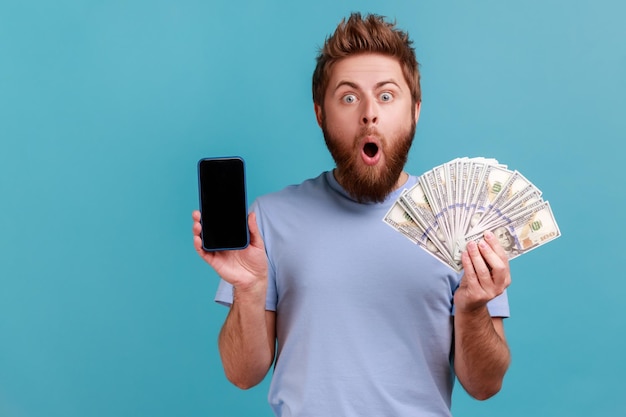 Man in blauwe t-shirt met dollarbankbiljetten en mobiele telefoon met leeg scherm online wedden