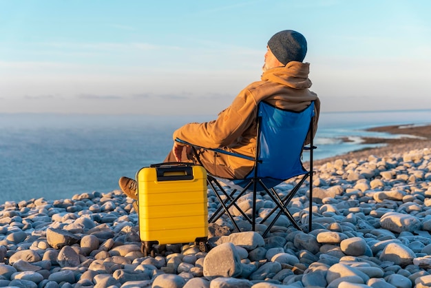 Man in bruine jas met gele koffer alleen ontspannen en zittend op strandstoelen aan zee