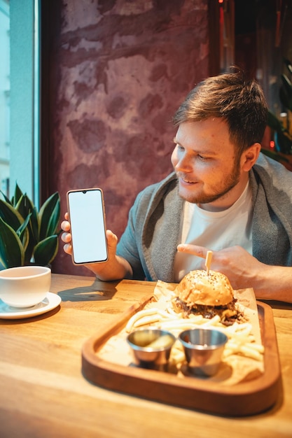 Man in café met telefoon met wit scherm die hamburger eet en thee drinkt