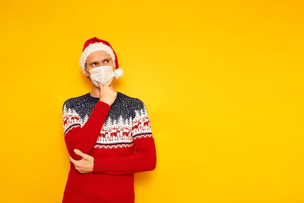 사진 순록 빨간 산타 클로스 모자 의료 마스크와 크리스마스 스웨터에 남자는 사려 깊은 포즈에 서