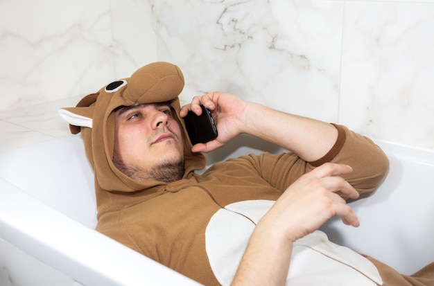 Man in cosplay kostuum van een koe. Kerel in de grappige dierenpyjama ligt in het bad en spreekt op de mobiele telefoon.