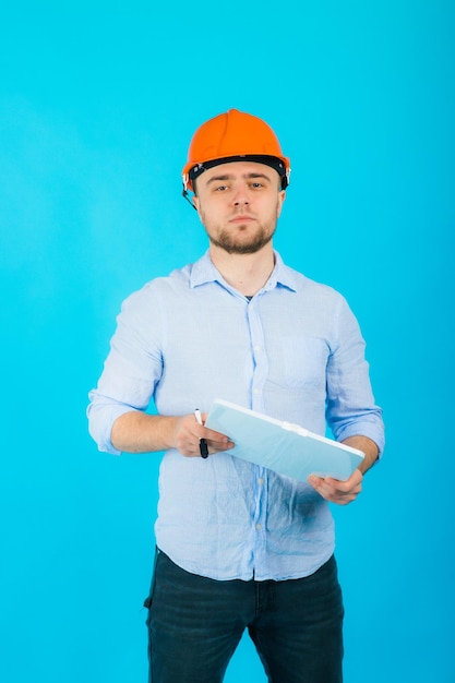 man in een blauw shirt in een beschermende oranje helm en een wit notitieboekje staat op een blauwe achtergrond