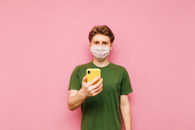 Man in een groen T-shirt en een wit medisch masker staat op een roze achtergrond met een smartphone in zijn hand kijkend naar de camera met een serieus gezicht Coronavirus Individuele bescherming in een pandemie