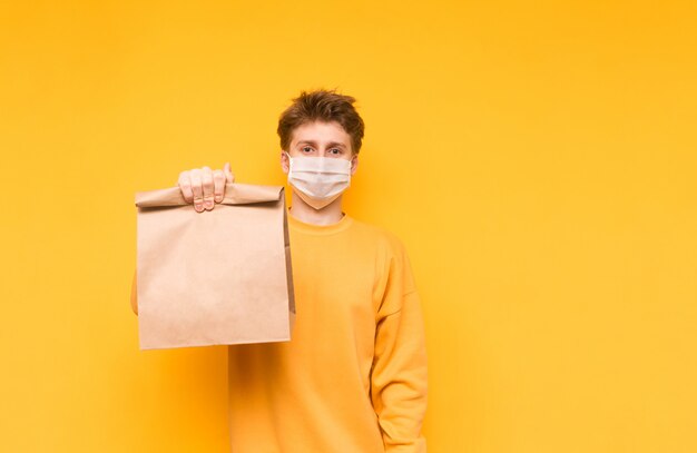 Man in een medisch masker en met een papieren zak poseert op een geel en biedt voedsel uit de bevalling. coronapandemie.