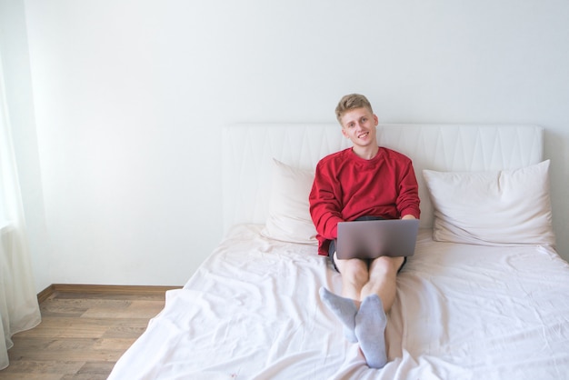 Man in een rode sweater zit op het bed in een lichte kamer en werkt op laptop