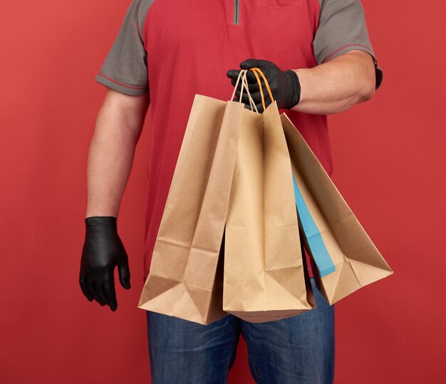 Man in een rood t-shirt, gekleed in zwarte latex handschoenen, houdt papieren zakken voor aankopen