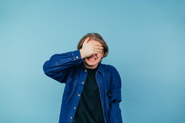 man in een shirt staat op een blauwe achtergrond, klemt zijn tanden op elkaar en bedekt zijn ogen van schaamte