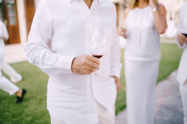 Man in een wit overhemd en broek staat met een glas rode wijn in zijn hand