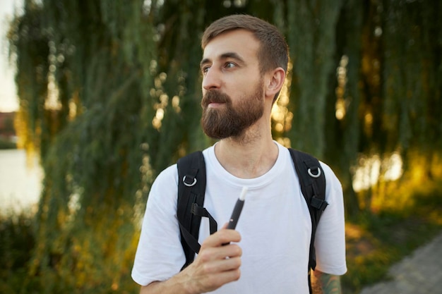 man in een wit t-shirt met een elektronische sigaret in het park. De man heeft een elektronische vast