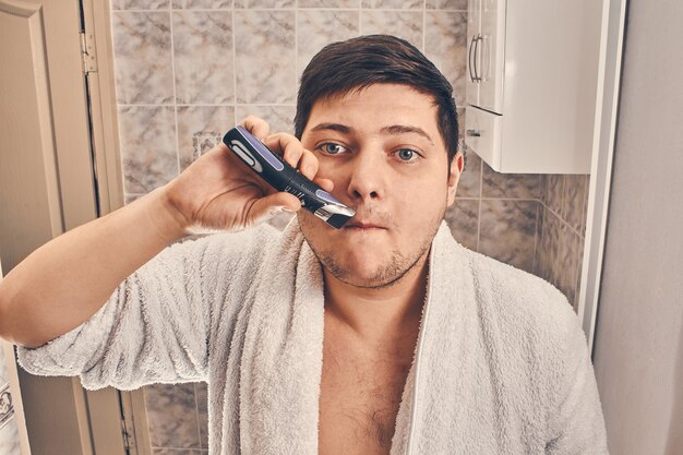 Man in een witte jas scheert zich met een trimmer in de badkamer.