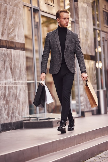 Man in elegante formele kleding en met winkelpakketten is buiten tegen modern gebouw.