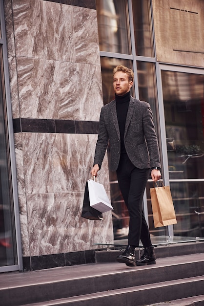 Man in elegante formele kleding en met winkelpakketten is buiten tegen modern gebouw.