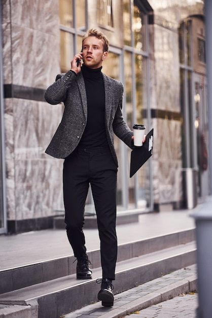 Man in elegante formele kleding met kopje drank en notitieblok in handen is buiten tegen modern gebouw en heeft een telefoongesprek.