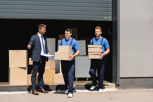 Man in formele kleding die klembord vasthoudt en met de hand wijst in de buurt van verhuizers met kartonnen dozen en