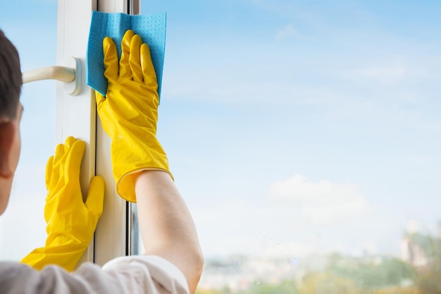 Man in gele handschoenen raam schoonmaken met doek en spray wasmiddel op huis terras Huis schoonmaken en huishoudelijke klusjes huishoudelijke hygiëne Glazenwassen achtergrond met blauwe lucht en kopieer ruimte