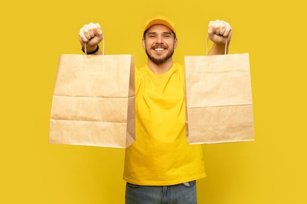 Man in gele pet, t-shirt met geïsoleerde papieren pakketten. Mannelijke werknemerskoerier houdt papieren pakjes met voedsel vast