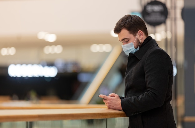 Man in gezichtsmasker en jas gebruikt mobiele telefoon in winkelcentrum in pandemische tijd