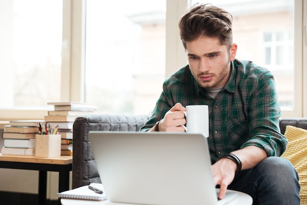 Man in groen shirt met kopje thee in de hand kijken naar laptop en zittend op de bank.