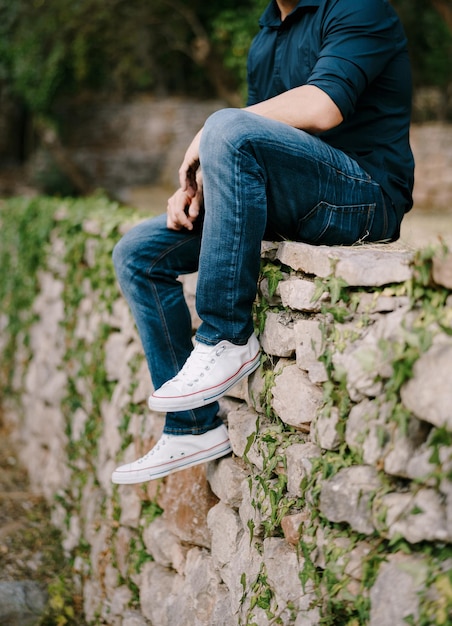 Man in jeans en sneakers zit op een stenen muur in de park-closeup