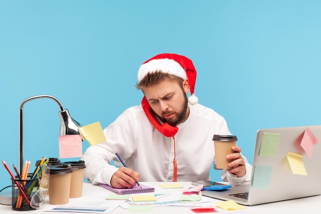 Man in Kerstman hoed zittend op de werkplek koffie drinken en praten vaste telefoon informatie opschrijven in Kladblok werken op vakantie Indoor studio shot geïsoleerd op blauwe achtergrond