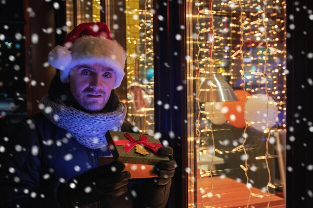 Man in kerstmuts met geschenkdoos in de buurt van verlicht caféraam