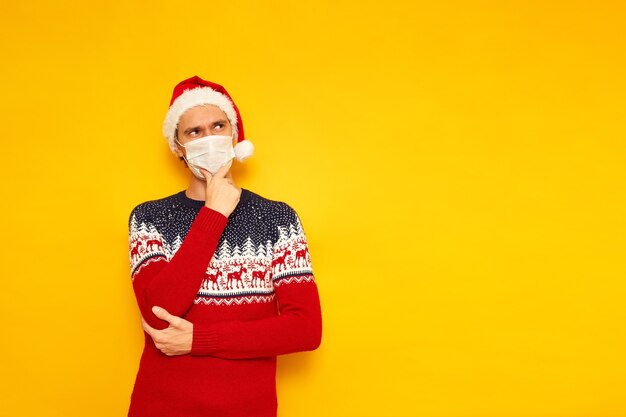 man in kersttrui met rendier rode kerstman hoed medisch masker staat in doordachte pose