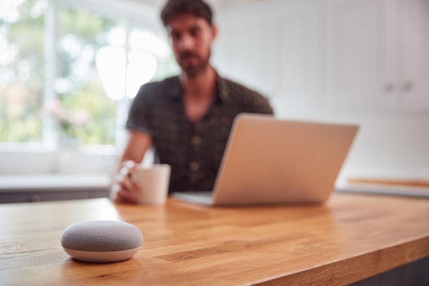 Man in keuken met behulp van digitale slimme luidspreker tijdens het werken vanuit huis met laptop tijdens pandemie