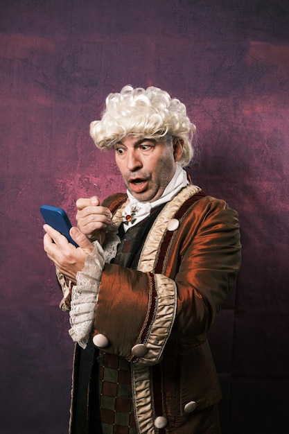 Foto man in klassieke renaissancekleding met een verbaasde uitdrukking op zijn gezicht terwijl hij naar het scherm van de mobiele telefoon kijkt met een monocle.