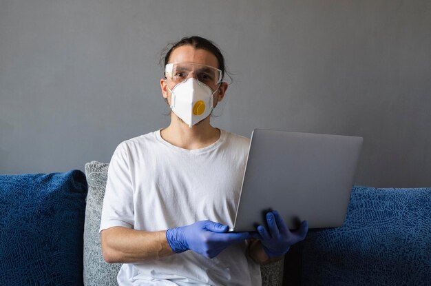 Man in medisch masker en rubberen handschoenen zit thuis en werkt met laptop op de bank tijdens quarantaine