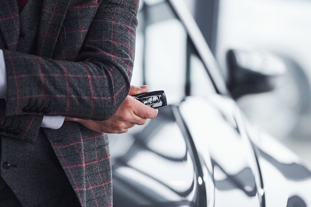 Man in moderne formele kleding staat tegen zwarte auto met sleutels in de hand.