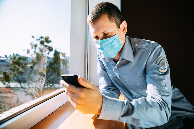 Man in quarantaine vanwege een virus zit thuis in een masker en kijkt uit het raam op een zonnige dag.