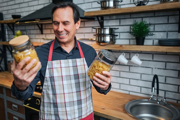 Man in schort staan in de keuken en houdt twee potten spagetti. Hij kijkt naar hen en glimlacht. Man staat alleen.