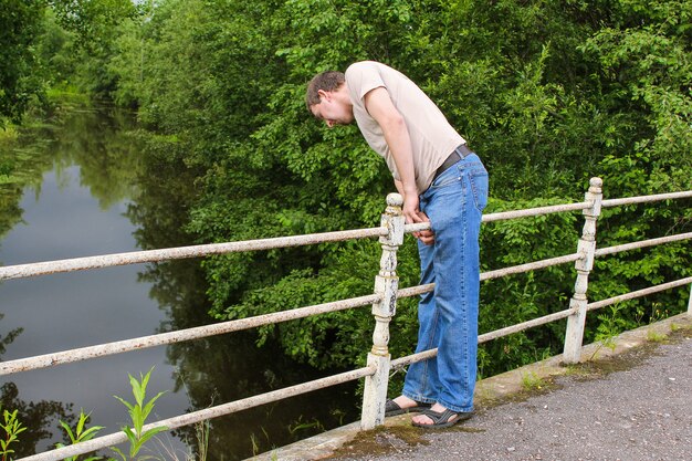 Man in spijkerbroek op brug die neerkijkt op het water tegen de achtergrond van groene bomen