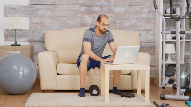 Man in sportkleding zoeken biceps training met halter op laptop zittend op de bank.