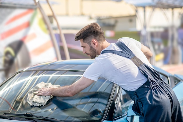 Man in t-shirt en overall die autoruit schoonveegt