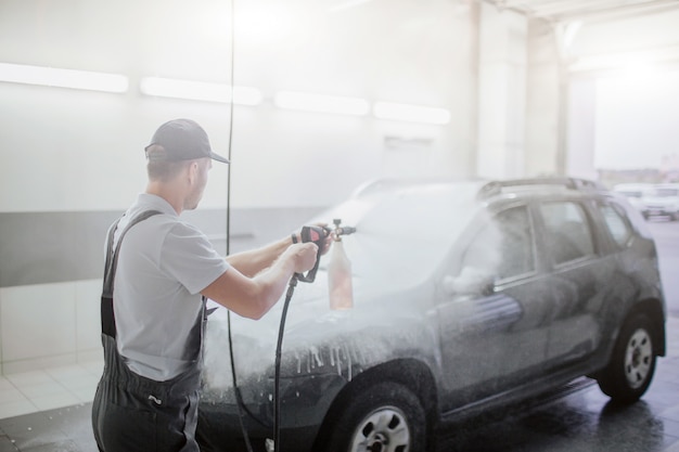 Man in uniform staat voor auto en wast het met water dat uit flexibele slang gaat. Zwarte auto is bedekt met schuim. Guy gaat het schoonmaken.