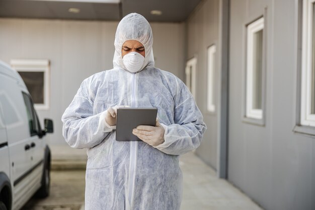 Man in virusbeschermend pak en masker kijkt en typen op tablet, gebouwen van coronavirus desinfecterend met de sproeier. epidemie.