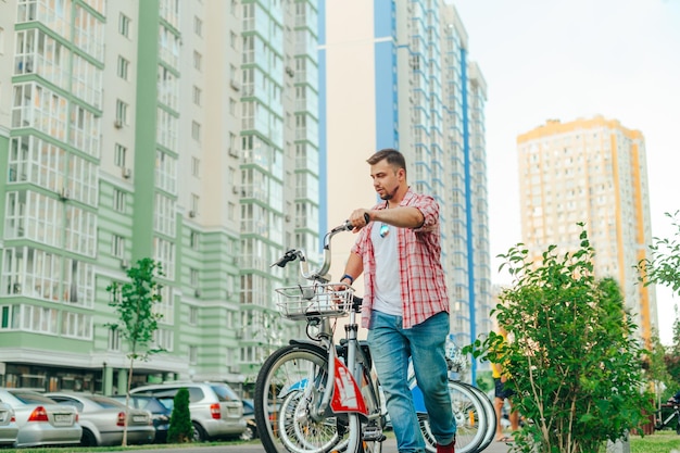 Man in vrijetijdskleding duwt een gehuurde stadsfiets tegen de achtergrond van een stedelijk landschap met appartementsgebouwen De toerist neemt een fiets naar de Schering voor een wandeling