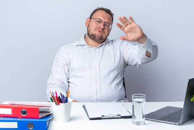 ラップトップのオフィスフォルダーとオフィスで働いている白い壁の上にクリップボードを持ってテーブルに座って手を振って真面目な顔で正面を見て眼鏡をかけている白いシャツの男