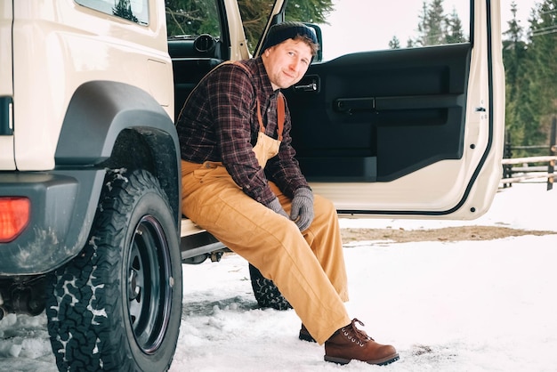 Man in winterkleren zit in de buurt van zijn auto en geniet van een besneeuwde dag in de buurt van bos