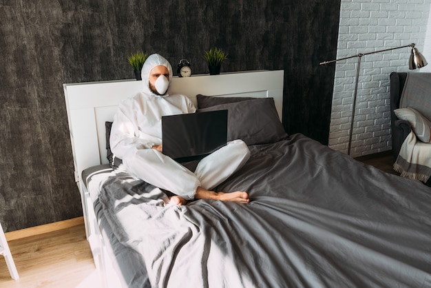 Man in wit beschermend kostuum met kap en gezichtsmasker thuis op het bed met laptop