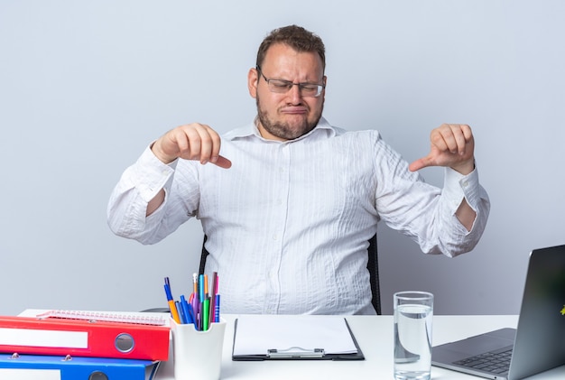 Man in wit overhemd met een bril die aan de tafel zit met kantoormappen en klembord die naar het scherm van de laptop kijkt en duimen naar beneden laat zien die ontevreden zijn over de witte muur die op kantoor werkt