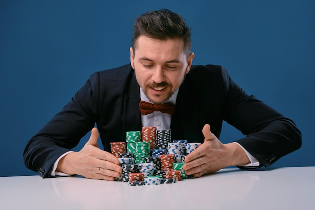 Man in zwart pak zit aan witte tafel met gekleurde stapels chips erop poseren op blauwe studio...