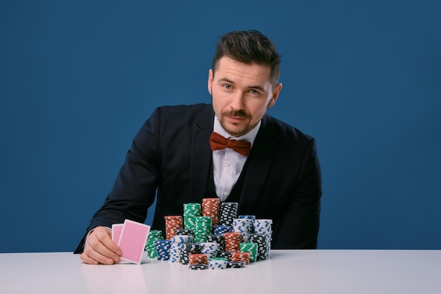 Man in zwart pak zit aan witte tafel met stapels chips met twee speelkaarten die poseren op...