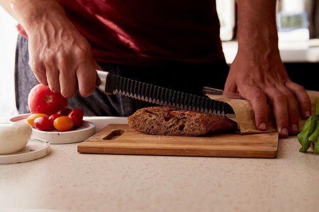 Man is cutting a French fresh grain baguette in the kitchen Basil tomatoes and mozzarella on the table Preparing a dinner