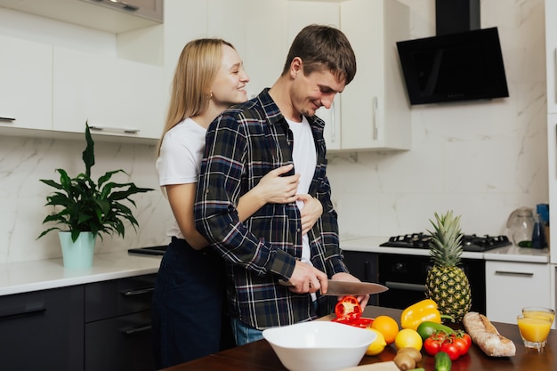 L'uomo sta tagliando le verdure in cucina e sorride mentre la sua donna lo abbraccia da dietro