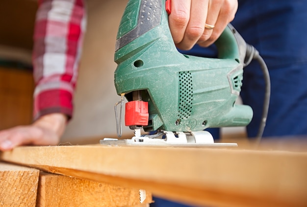 The man is cutting the wooden plank by jigsaw