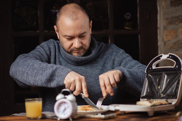 The man is having breakfast An ordinary middleaged man in a sweater is preparing to eat a sandwich in a dark room